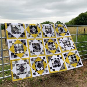Puede incluir: Un edredón hecho a mano con un patrón geométrico. El edredón presenta bloques cuadrados con patrones de cuadros en blanco, negro y gris, enmarcados por bordes amarillos y blancos. El edredón está colgado sobre una valla metálica al aire libre.