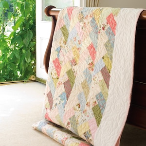 May include: A patchwork quilt with a diamond pattern in pastel colors, including pink, blue, yellow, and green. The quilt is draped over a wooden headboard, with a matching pillow at the base. The quilt has a white border with a floral pattern.