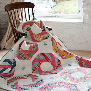 Op de afbeelding: Een kleurrijk quilt met een geometrisch patroon in roze, blauw, geel en groen. De quilt is gemaakt van stofresten en is gevouwen over een houten schommelstoel.