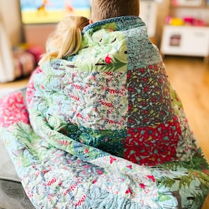 May include: Two children are snuggled under a colorful patchwork quilt while watching television. The quilt is made of various fabrics with floral and woodland patterns in shades of green, blue, red, and white.