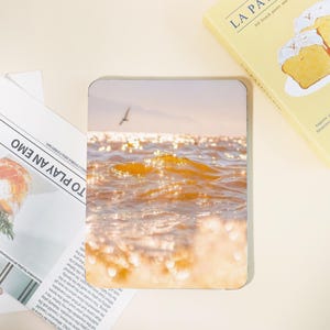 Könnte beinhalten: Eine rechteckige Handyhülle mit einer ruhigen Meereslandschaft, in der sich goldenes Sonnenlicht auf dem Wasser spiegelt. Eine Möwe fliegt in der Ferne. Die Hülle liegt neben einer Zeitung mit dem Text "TO PLAY AN EMO" und einem Buch mit dem Titel "LA PA".