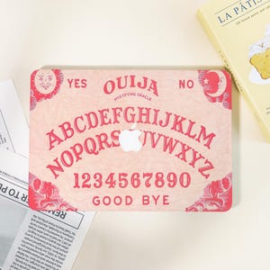 May include: A pink laptop skin featuring a Ouija board design with red lettering and graphics. The design includes the alphabet, numbers, and the words "YES", "NO", and "GOOD BYE". A white Apple logo is in the centre.