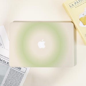 May include: A laptop with a gradient green and white design. The Apple logo is visible in the center. A book titled "La Pâtisserie" and a newspaper are in the background.