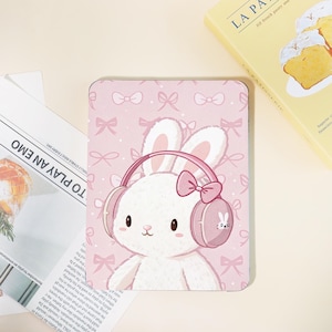 May include: A pink tablet case featuring a cartoon bunny wearing pink headphones with a bow. The background is a light pink with bow patterns. A book with the text "LA PA" is in the background, along with a newspaper with the text "TO PLAY AN EMO".