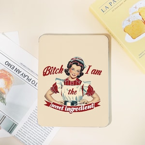 May include: A cream-colored metal sign featuring a vintage illustration of a woman in a red and white apron. The text reads "Bitch I am the secret ingredient" in red and black.