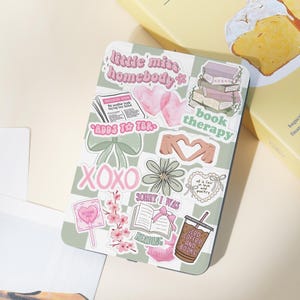 Könnte beinhalten: Eine Tablet-Hülle mit grün-weißem Karomuster, verziert mit verschiedenen Aufklebern. Die Aufkleber zeigen Texte wie "little miss homebody" und "book therapy". Weitere Aufkleber sind Herzen, Schleifen, Blumen und ein Stapel Bücher.