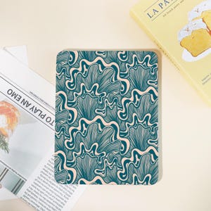 May include: A rectangular e-reader device featuring a dark teal and light peach abstract pattern of wavy, fan-like lines. The device is positioned on a flat surface next to a white paper with printed text and a yellow book with a pastry illustration.