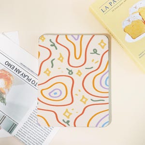 May include: A rectangular cream-colored notepad with a colorful abstract design of wavy lines, stars, and confetti-like shapes. The notepad is next to a newspaper with the headline "TO PLAY AN EMO" and a yellow box with the text "LA PA".