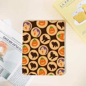 May include: A rectangular Halloween-themed item featuring a pattern of round cookies. Each cookie has a brown base with orange pumpkin, black cat, and purple ghost designs. The background is dark brown.