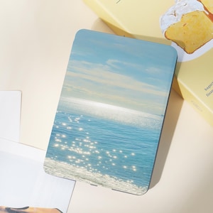 May include: A rectangular, blue and white decorative item featuring a seascape with a bright sun reflecting on the water. The image shows a clear sky and sparkling water. The item is placed on a light-colored surface.