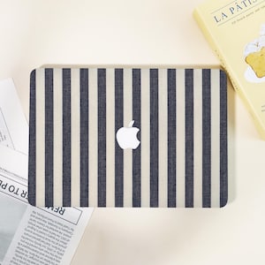 May include: A laptop case with a navy blue and off-white vertical striped pattern. The Apple logo is centered on the case. A book titled "La Pâtisserie" and a newspaper are in the background.