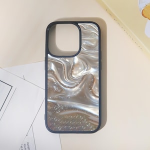 May include: A smartphone case with a pearlescent, swirling design in shades of silver and white. The case has a dark blue border and a cutout for the camera. The bottom of the case has a glittery effect.