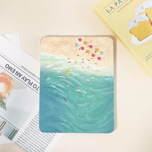 May include: A rectangular art print depicts a beach scene with turquoise water and a sandy shore. Colorful umbrellas dot the beach, and small figures are in the water. A yellow book with the text "LA PA" is in the background.