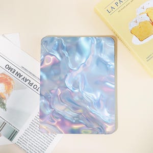 May include: A rectangular, iridescent case with a flowing, liquid-like design in shades of blue, purple, and silver. The case has rounded corners and a glossy finish. The text "TO PLAY AN EMO" is visible on a newspaper.