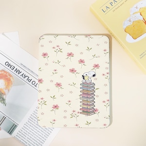 Könnte beinhalten: Eine rechteckige E-Reader-Hülle mit cremefarbenem Hintergrund und einem wiederkehrenden Muster aus kleinen rosa Blüten und grünen Blättern. Ein Zeichentrickhund sitzt auf einem hohen Stapel bunter Bücher und liest ein kleines Buch.