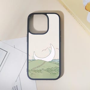 May include: A phone case with a navy blue border and a cream-colored background. The case features a minimalist illustration of a crescent moon over rolling green hills, with a path leading towards it. The design has a whimsical, artistic feel.