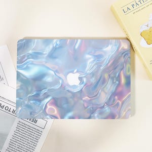 May include: A laptop case with a pearlescent, iridescent design in shades of blue, purple, and silver. The Apple logo is visible in white. The case has a glossy finish and a flowing, liquid-like pattern.