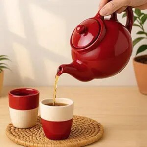 Könnte beinhalten: Eine rote Keramik-Teekanne gießt Tee in eine rot-cremefarbene Tasse. Eine weitere Tasse steht daneben auf einem gewebten Untersetzer. Die Teekanne und die Tassen stehen auf einer hellbraunen Oberfläche, mit einer Pflanze im Hintergrund.