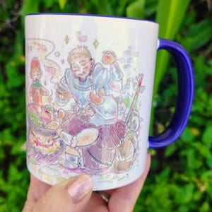 May include: White ceramic mug with a blue interior and handle. The mug features a colourful illustration of cartoon characters in a medieval setting, with a focus on cooking. The artwork includes a variety of colours and details.