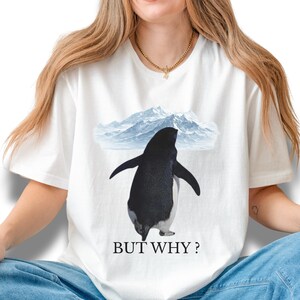 Puede incluir: Camiseta blanca con un diseño de pingüino en blanco y negro con las alas extendidas, sobre un fondo de montañas nevadas. El texto "BUT WHY?" está impreso debajo.