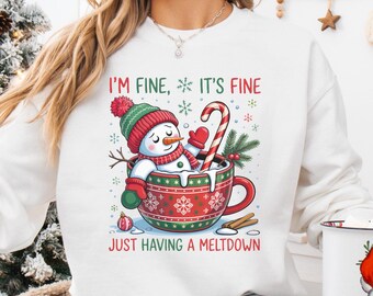 Può includere: Felpa bianca con una grafica di un pupazzo di neve in una tazza rossa e verde, con la scritta "I'M FINE, IT'S FINE JUST HAVING A MELTDOWN". Il pupazzo di neve indossa un cappello, una sciarpa e guanti rossi, con un bastoncino di zucchero nella tazza.