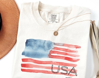 May include: Cream-colored t-shirt featuring a watercolor American flag design in red and blue. The word "USA" and the number "1776" are printed in black. A gold necklace and a brown hat are also visible.