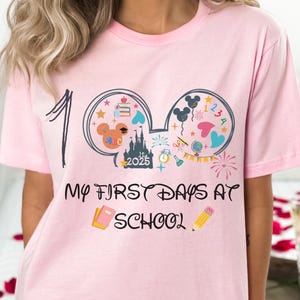 May include: Pink t-shirt with a graphic design featuring Mickey Mouse ears filled with school-themed illustrations. The shirt has the text "100" and "My First Days at School" in a playful font. A pencil and a notebook are also featured.