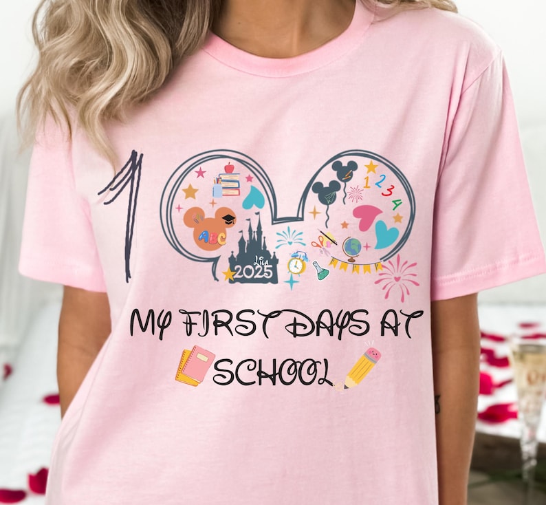 May include: Pink t-shirt with a graphic design featuring Mickey Mouse ears filled with school-themed illustrations. The shirt has the text "100" and "My First Days at School" in a playful font. A pencil and a notebook are also featured.