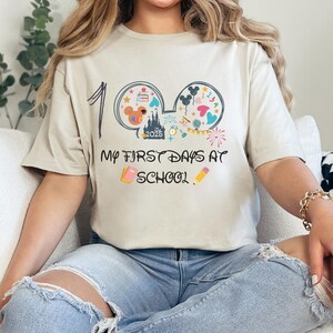 May include: Beige t-shirt with the text "My First Days at School" and a graphic of Mickey Mouse ears filled with school-themed icons. The number "1" is written above the ears. The shirt is worn by a person.