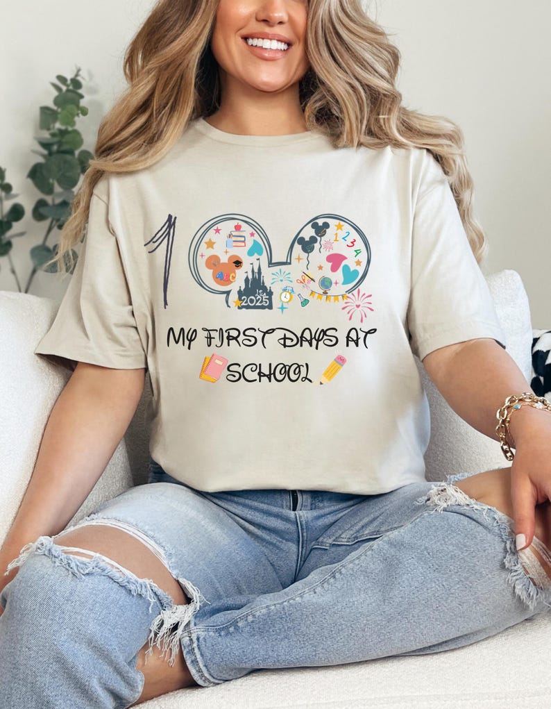 May include: Beige t-shirt with the text "My First Days at School" and a graphic of Mickey Mouse ears filled with school-themed icons. The number "1" is written above the ears. The shirt is worn by a person.