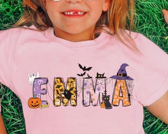 Może przedstawiać: Różowa koszulka z napisem "EMMA" w halloweenowym motywie. Litery ozdobione są nietoperzami, kapeluszem czarownicy, czarnym kotem, dynią i duchem. Koszulkę nosi dziecko w białych okularach przeciwsłonecznych.