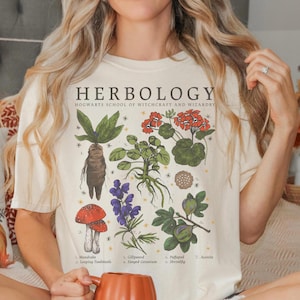 May include: Cream-colored t-shirt featuring a botanical illustration with the text "HERBOLOGY" and "HOGWARTS SCHOOL OF WITCHCRAFT AND WIZARDRY". The design includes drawings of various plants, including a mandrake and toadstools.