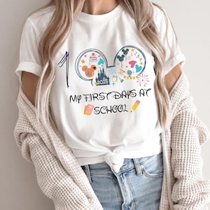 May include: White t-shirt with a colorful graphic design featuring Mickey Mouse ears, a castle, and the text "My First Days at School 2025." The design includes a number 1, a book, and a pencil.