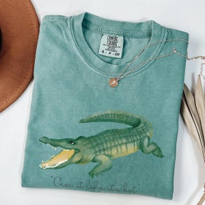 Könnte beinhalten: Ein teal Comfort Colors T-Shirt mit einer grünen Alligator-Illustration und dem Schriftzug "Croc it like it's hot". Eine silberne Halskette mit Anhänger liegt über dem Shirt.