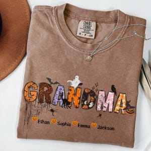 May include: A brown Comfort Colors t-shirt with the word "GRANDMA" in a Halloween-themed design. The letters are decorated with bats, spiders, pumpkins, and ghosts. Below the word are the names Ethan, Sophia, Emma, and Jackson.