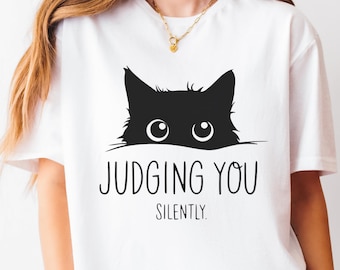 May include: White t-shirt featuring a black cat graphic with the text "JUDGING YOU SILENTLY." The cat's eyes are visible above the text. The shirt is a casual, everyday wear item.