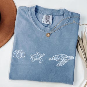 May include: A blue Comfort Colors t-shirt with a white turtle design. The shirt has a crew neck and short sleeves. A gold necklace with a pendant is draped across the shirt. The shirt is size small.