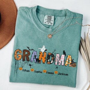 May include: A teal Comfort Colors t-shirt with the word "GRANDMA" in a Halloween-themed design. The letters are decorated with bats, spiders, and pumpkins. Below the word are the names Ethan, Sophia, Emma, and Jackson.