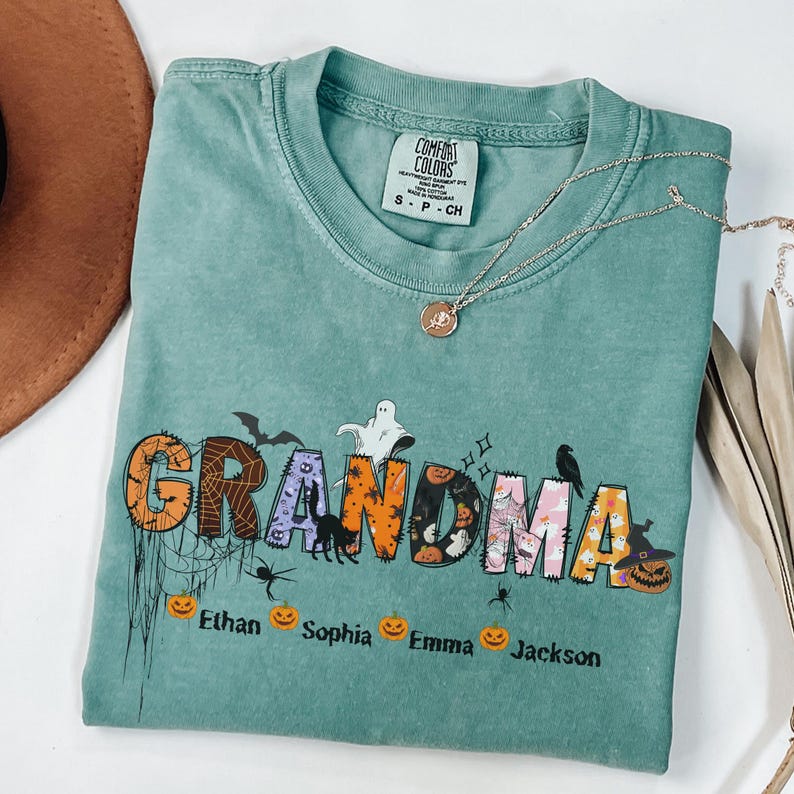 May include: A teal Comfort Colors t-shirt with the word "GRANDMA" in a Halloween-themed design. The letters are decorated with bats, spiders, and pumpkins. Below the word are the names Ethan, Sophia, Emma, and Jackson.