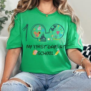 May include: A green t-shirt with the text "My First Days at School" and a graphic of Mickey Mouse ears filled with school-themed icons. The number "1" is on the left side of the shirt. A pencil and book are also featured.