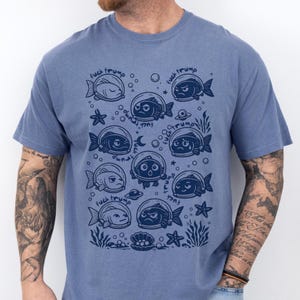 Op de afbeelding: Een blauw t-shirt met een grillig ontwerp van vissen met astronautenhelmen. De vissen hebben verschillende gezichtsuitdrukkingen en zijn omringd door bellen, sterren en de tekst "fuck trump".