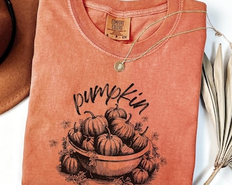Op de afbeelding: Oranje Comfort Colors T-shirt met een zwarte afbeelding van pompoenen in een kom en het woord "pumpkin" erboven. Een gouden ketting en een bruine hoed zijn ook zichtbaar.