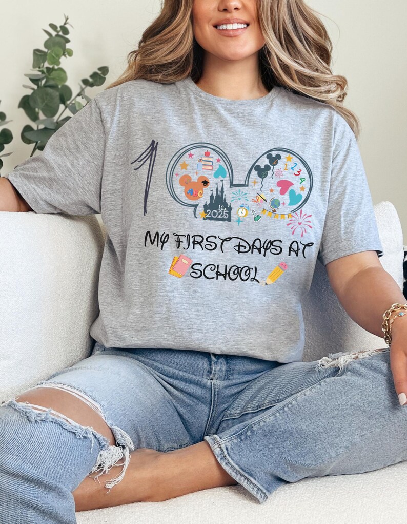May include: A gray t-shirt with a colorful graphic design featuring Mickey Mouse ears, a castle, and the text "My First Days at School." The shirt also includes the number "1" and the year "2025."