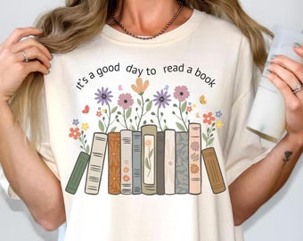 Puede incluir: Camiseta color crema con la frase "It's a good day to read a book" sobre una fila de libros y flores coloridas. Los libros tienen varios diseños y colores, con mariposas dispersas alrededor de las flores.