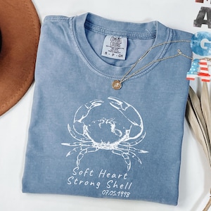 Peut inclure: Un t-shirt bleu Comfort Colors avec un motif de crabe blanc et le texte "Soft Heart Strong Shell 07.05.1998." Un collier en or avec un pendentif est drapé sur le t-shirt.
