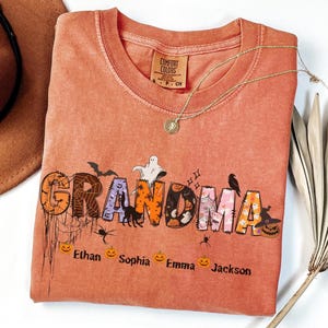 May include: A coral-colored Comfort Colors t-shirt with the word "GRANDMA" in a Halloween-themed design. The design includes bats, spiders, pumpkins, and a ghost. The names Ethan, Sophia, Emma, and Jackson are below the word.