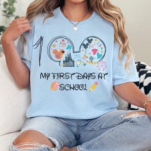 May include: Light blue t-shirt with a graphic featuring Mickey Mouse ears, a castle, and the text "My First Days at School." The design includes colorful elements like stars, balloons, and a pencil. The number "1" is printed on the left side.