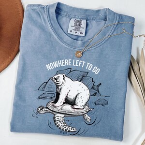 May include: A blue Comfort Colors t-shirt with a graphic of a polar bear on a turtle, and the text "NOWHERE LEFT TO GO." A gold necklace with a pendant is draped across the shirt.