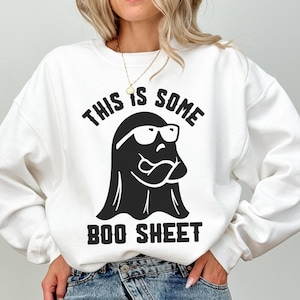 May include: White sweatshirt with a black graphic of a ghost wearing glasses and the text "THIS IS SOME BOO SHEET". The ghost has a mischievous expression. A fun, casual top for Halloween or any time.