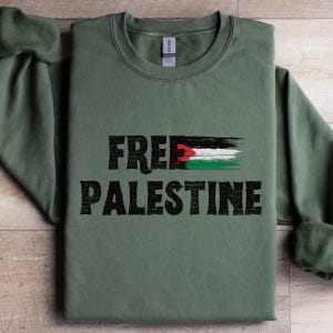 May include: Olive green sweatshirt with the words "FREE PALESTINE" in black, with a Palestinian flag design. The sweatshirt has a crew neck and long sleeves. The text and flag are printed on the front.